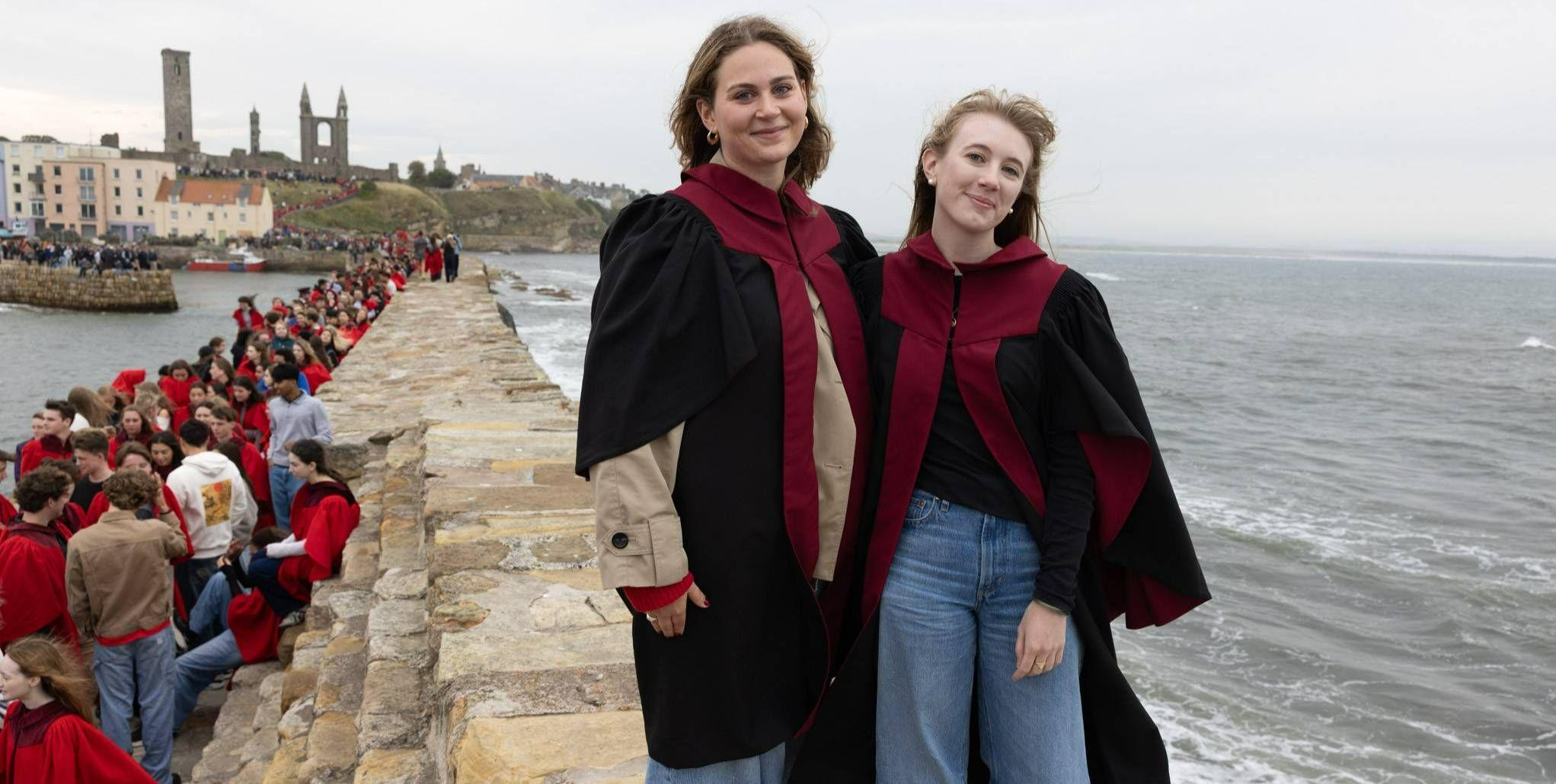 Two students stand smiling for a picture on the top of the pier in St Andrews. Waves are crashing to one side.