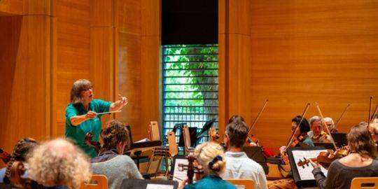 Jill conducts an orchestra in the McPherson Recital Room in the Laidlaw Music Centre. The musicians are visible but mainly with their backs to the camera - Jill is position to the left, waving her arms to conduct. Jill conducts an orchestra in the McPherson Recital Room in the Laidlaw Music Centre. The musicians are visible but mainly with their backs to the camera - Jill is position to the left, waving her arms to conduct.