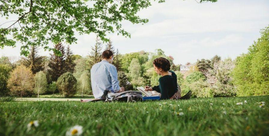 Image taken from behind Ewan and Karlee, they can be seen to be sitting on the grass on Hallow Hill. Karlee is sketching in a notebook - the background is full of trees with green leaves.