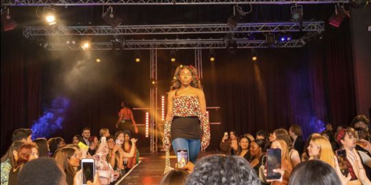 A student walks the runway in a previous Ubuntu fashion show, a crowd of students surround the catwalk, some taking pictures on their phones. 