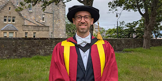 A university academic, Dr Adam Bower, stands on a lawn wearing a red and yellow formal academic robe and black cap with a gold tassel. A university academic, Dr Adam Bower, stands on a lawn wearing a red and yellow formal academic robe and black cap with a gold tassel.