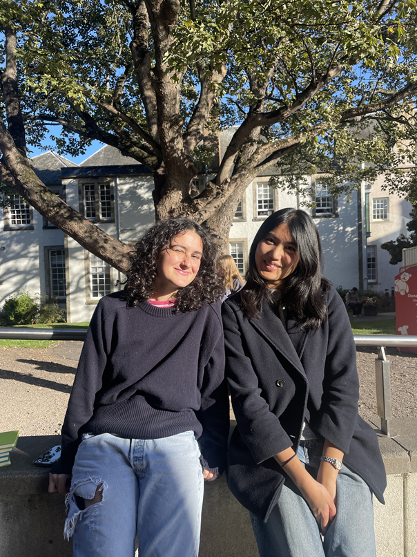 Two friends, both university students, perching on a wall in front of a tree. Blue sky is visible through the leaves. Two friends, both university students, perching on a wall in front of a tree. Blue sky is visible through the leaves.