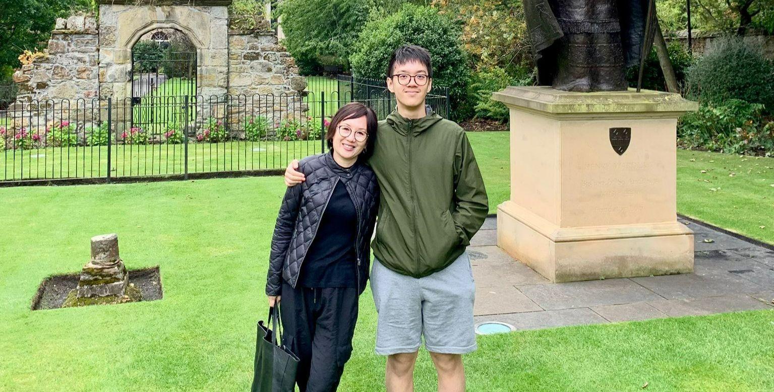 Two people - Helena Hu and her son - are standing on a green lawn in front of a historic stone structure and the plinth of a statue. Two people - Helena Hu and her son - are standing on a green lawn in front of a historic stone structure and the plinth of a statue.