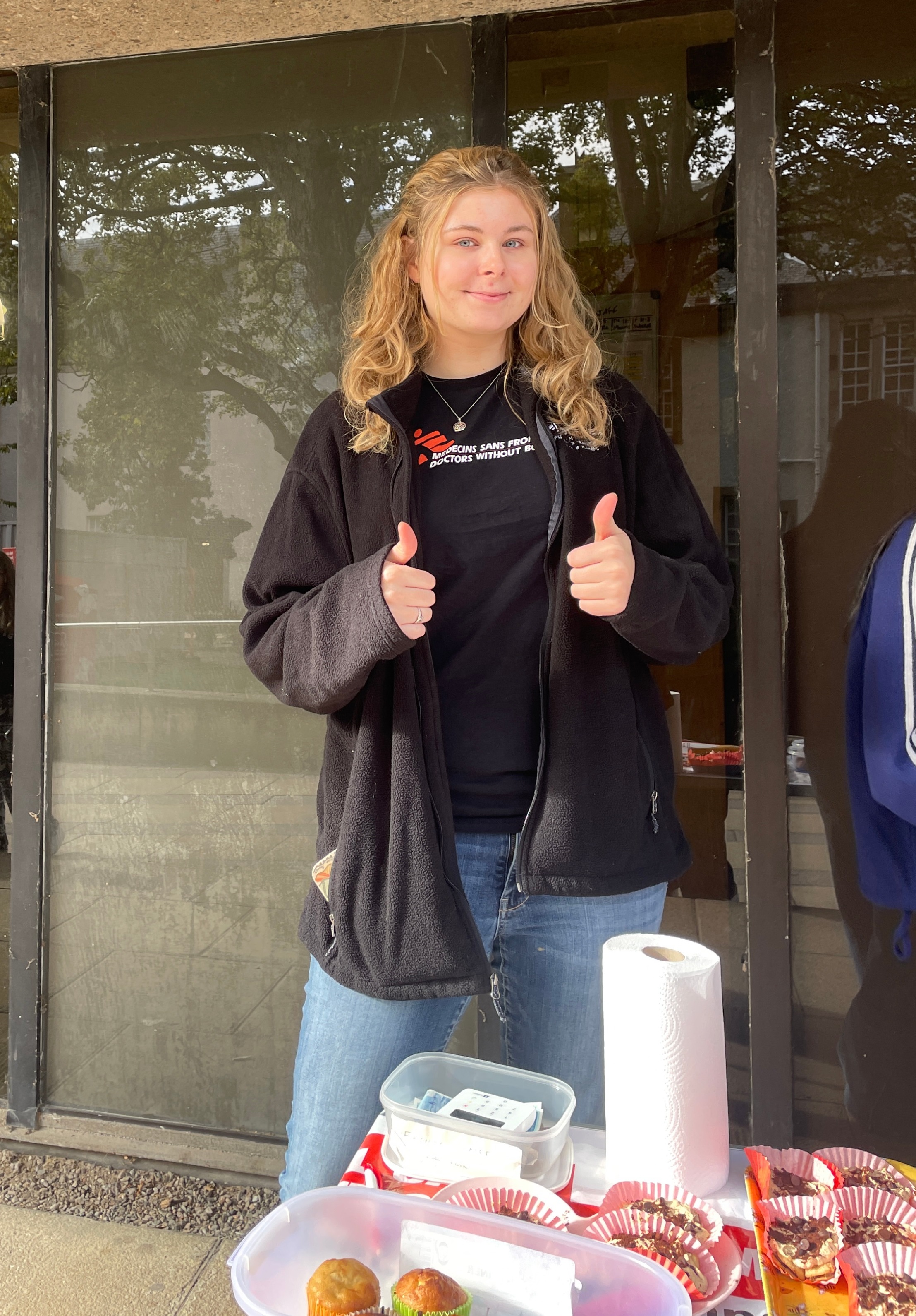 A student with blonde hair giving two thumbs up to the camera. She is standing behind a bake sale table with cakes and a red cloth. A student with blonde hair giving two thumbs up to the camera. She is standing behind a bake sale table with cakes and a red cloth.