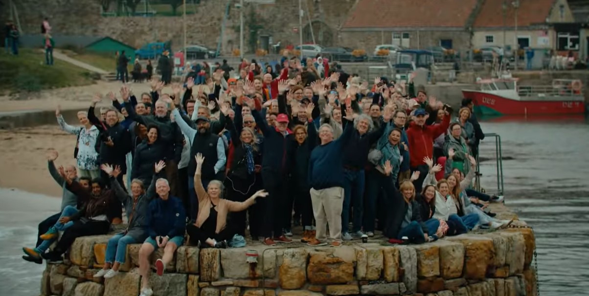 A large group of university students' parents are gathered seated and standing on the end of a pier. All are waving and cheering towards the camera. A large group of university students' parents are gathered seated and standing on the end of a pier. All are waving and cheering towards the camera.
