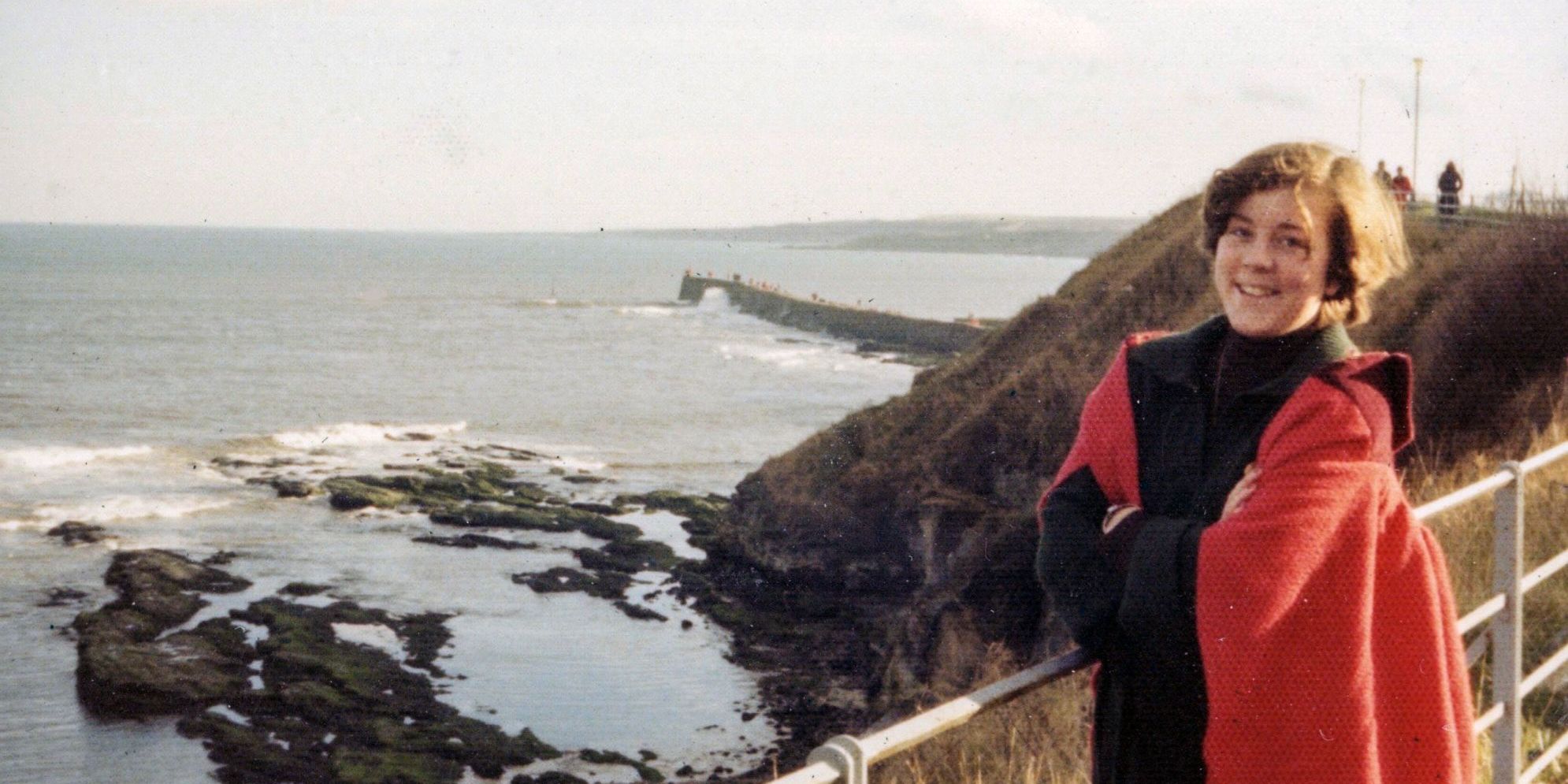 A female student in a red gown is pictured by the sea and cliffs in ���ϳԹ���. The photo dates from the late 1970s.