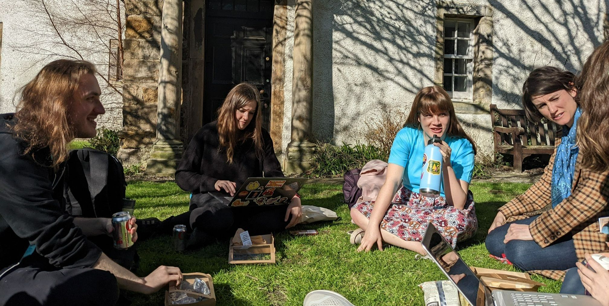 Students seated on the grass with books, laptops and packed lunch items