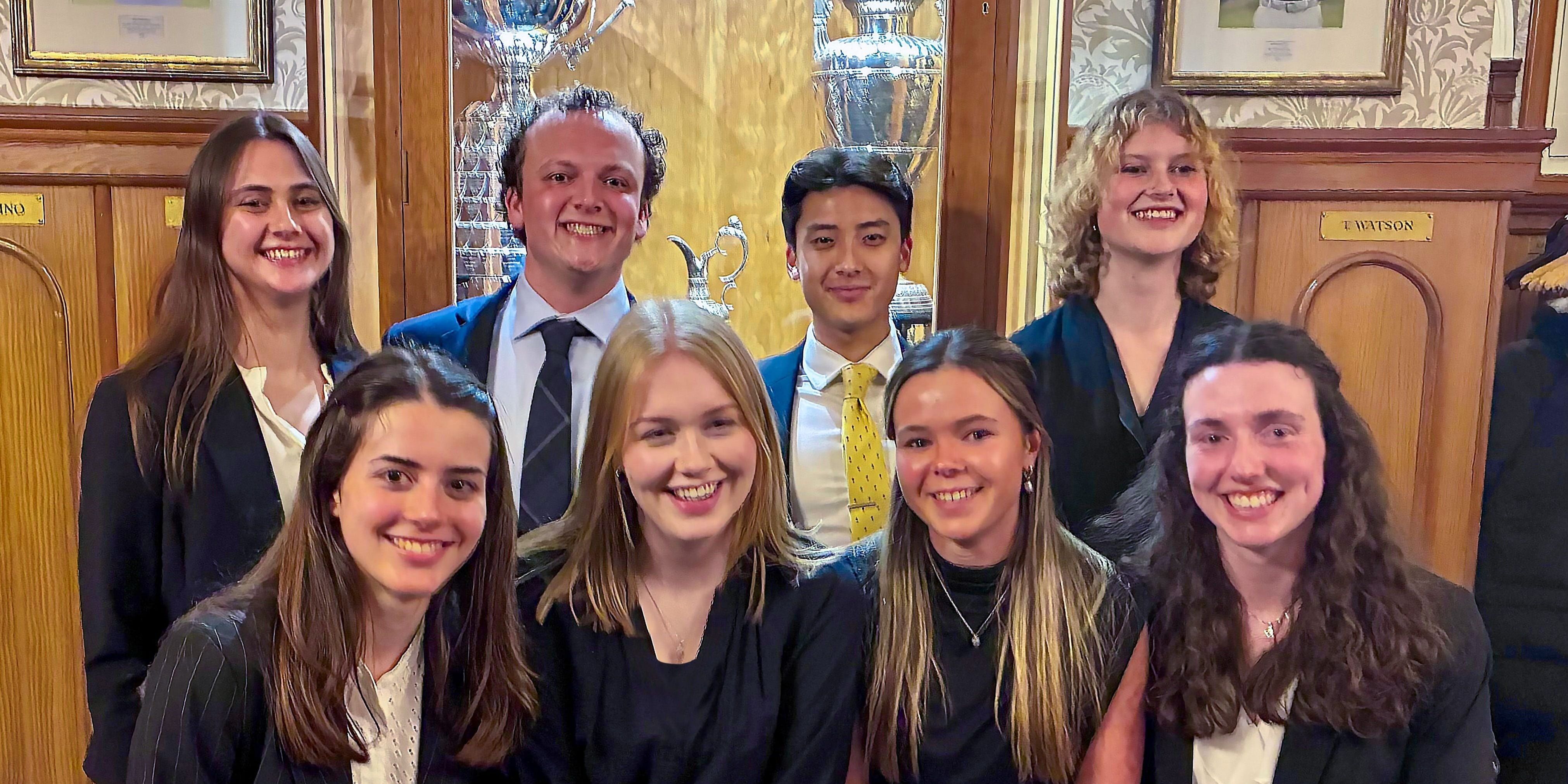 Eight Bobby Jones Scholars pose smiling for a photo at the R&A, a trophy cabinet can be seen in the background of the image
