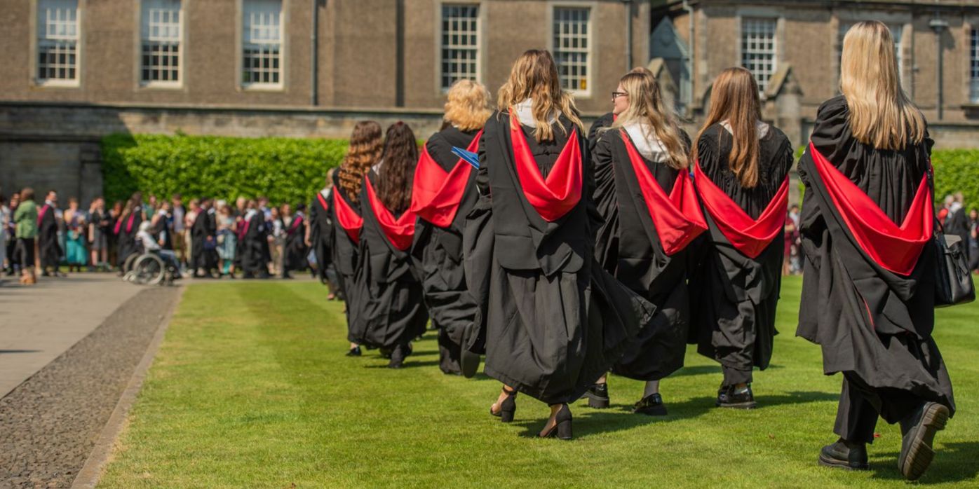 Graduates walking away from the camera in black gowns with red hoods. They are supported at the side of the lawn by a crowd of friends and relatives.
