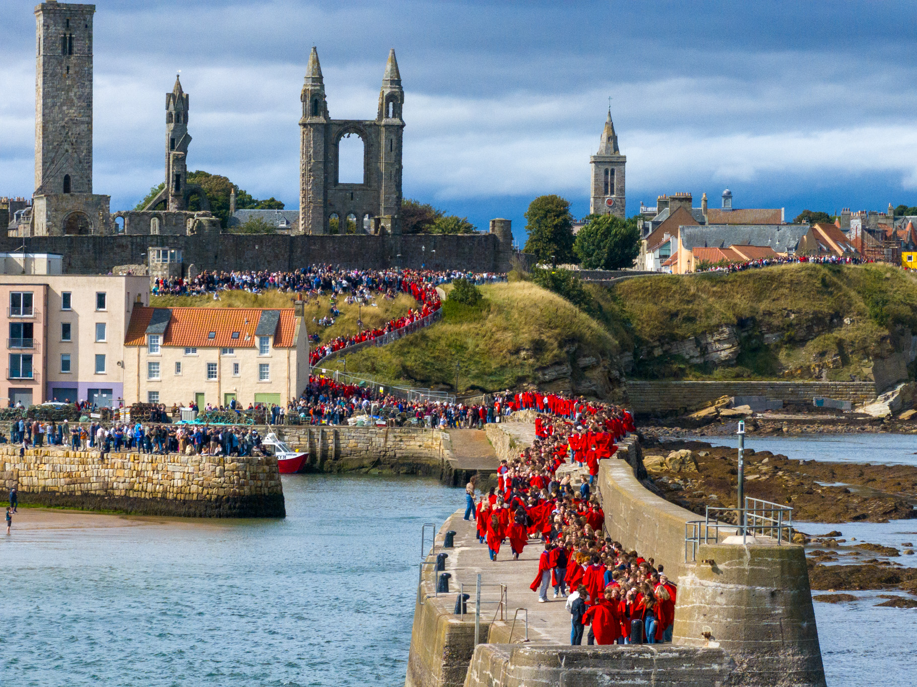 Crowds of students in red gowns gather on the path down and along the pier. The St Andrews skyline of the Cathedral can be seen in the background. Crowds of students in red gowns gather on the path down and along the pier. The St Andrews skyline of the Cathedral can be seen in the background.