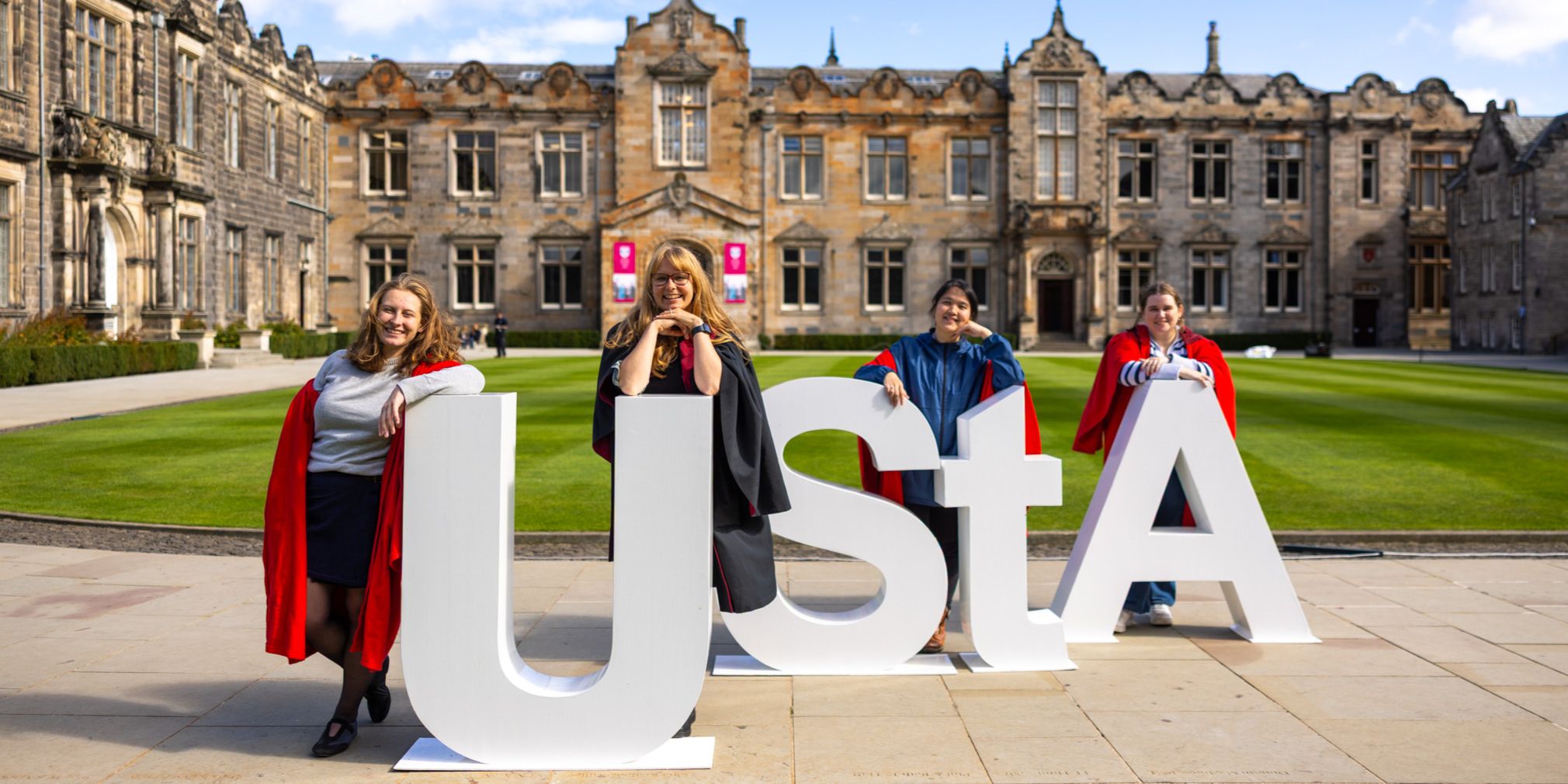 Students standing in St Salvator's Quad in red gowns lean against large white letters that read ' UStA' Students standing in St Salvator's Quad in red gowns lean against large white letters that read ' UStA'