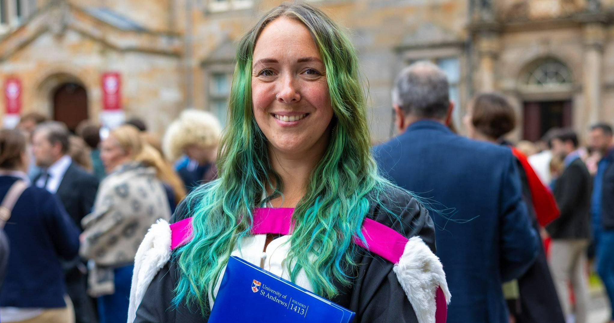 The image shows a female student graduating from the University of St Andrews. She is wearing a gown and hood and is holding a blue degree folder. She has long, blue hair. The image shows a female student graduating from the University of St Andrews. She is wearing a gown and hood and is holding a blue degree folder. She has long, blue hair.