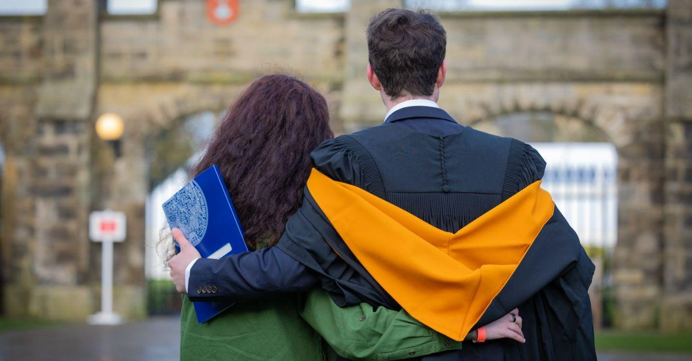 A student in a graduation gown and hood with his arm round a woman, looking away from the camera