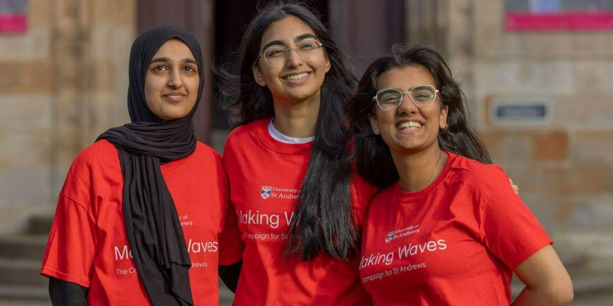 Three female students wearing red t-shirts with the Making Waves Campaign and University of St Andrews logos on the front, smiling and looking upwards.