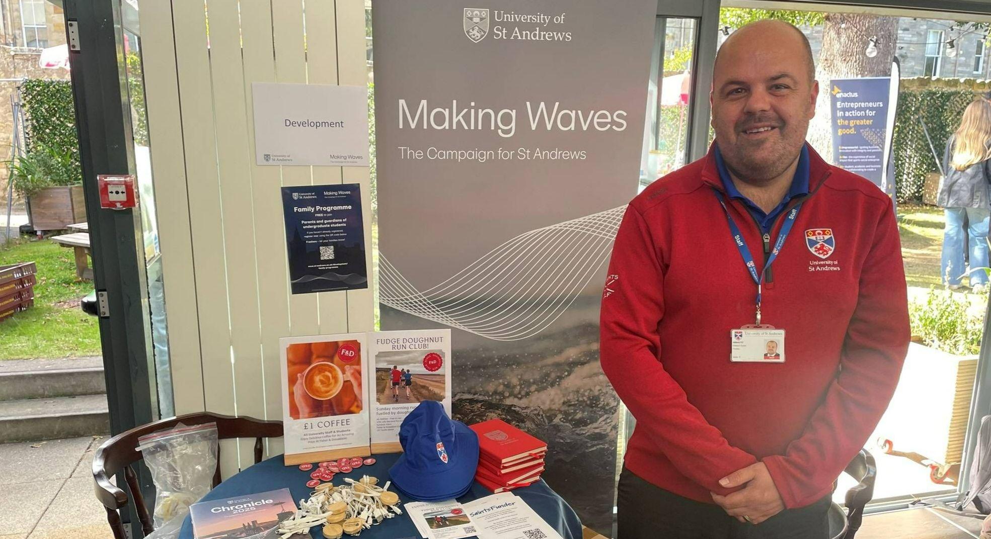 A male member of the Development team is pictured smiling, in front of the Development stand at freshers' fayre. A Making Waves pop up banner can be seen in the background, with a table full of St Andrews merchandise at the front.  