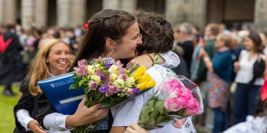 Two graduates hug in St Salvator's Quad, they both hold bouquets of flowers