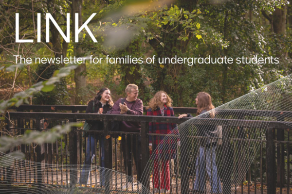 Four people standing on a bridge with black railings in a forested area, surrounded by lush green trees. The leaves are turning orange for autumn. 