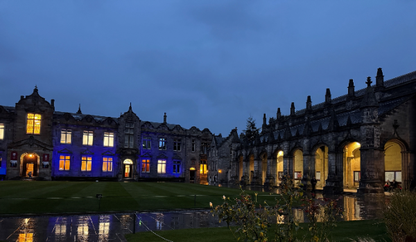 The University of St Andrews quad pictured on a dark autumn night in the rain. The historic buildings are lit blue and yellow.